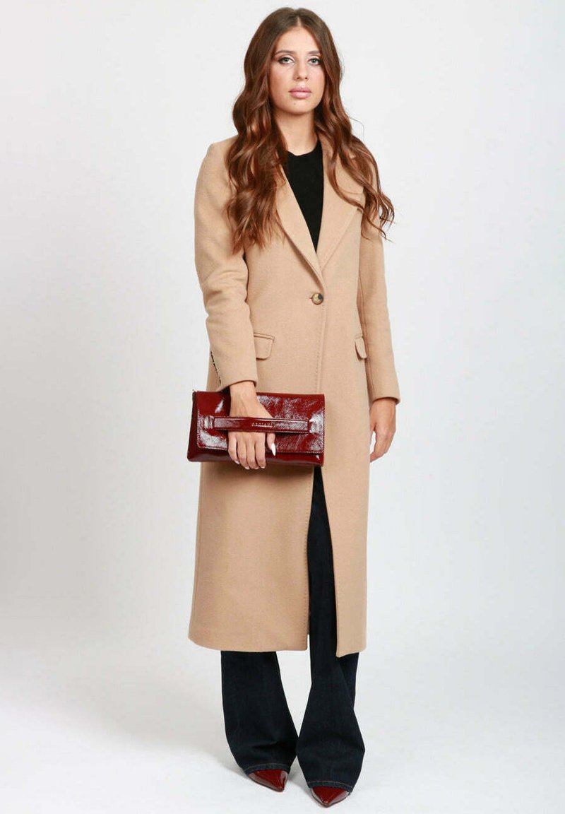 Donna con capelli lunghi castani, che indossa un lungo cappotto beige, pantaloni neri, scarpe a punta bordeaux e tiene in mano una brillante pochette bordeaux.