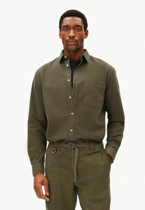Mann trägt ein olivgrünes Hemd mit Knöpfen und Brusttasche sowie passende Hose mit Gürtel, steht mit einer Hand in der Tasche vor einfarbigem Hintergrund.