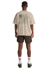 Homme portant un t-shirt graphique beige, un short foncé, des chaussettes blanches avec le texte « OFF DUTY GEAR » et des baskets noires, debout de dos sur fond blanc.
