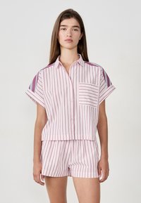 Femme portant une chemise à manches courtes, boutonnée, rayée rose avec un short assorti, se tenant devant un fond blanc uni.