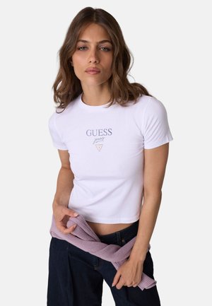 Donna con capelli castani ondulati indossa una maglietta bianca aderente Guess jeans e tiene un maglione lavanda piegato intorno alla vita.