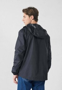 Zwarte waterdichte jas met capuchon, voorzien van elastische manchetten en een rechte snit. Draagt over blauwe spijkerbroek en heeft een gladde textuur.