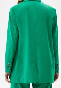 Blazer vert à coupe ajustée, avec un col cranté, une texture lisse et trois boutons décoratifs sur chaque poignet de manche.