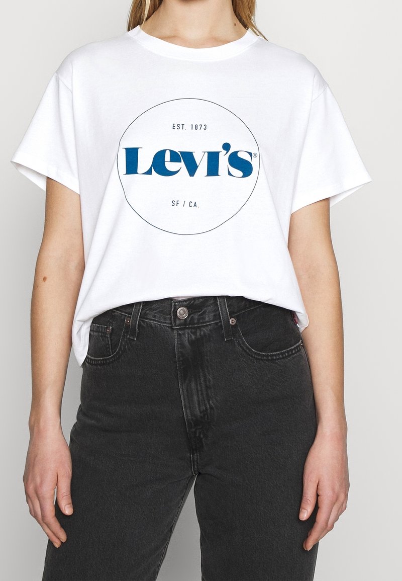 Bílý bavlněný tričko s modrým logem "Levi's" v kruhovém designu, nošeno s černými kalhotami s vysokým pasem, které mají zapínání na knoflík a kapsy.