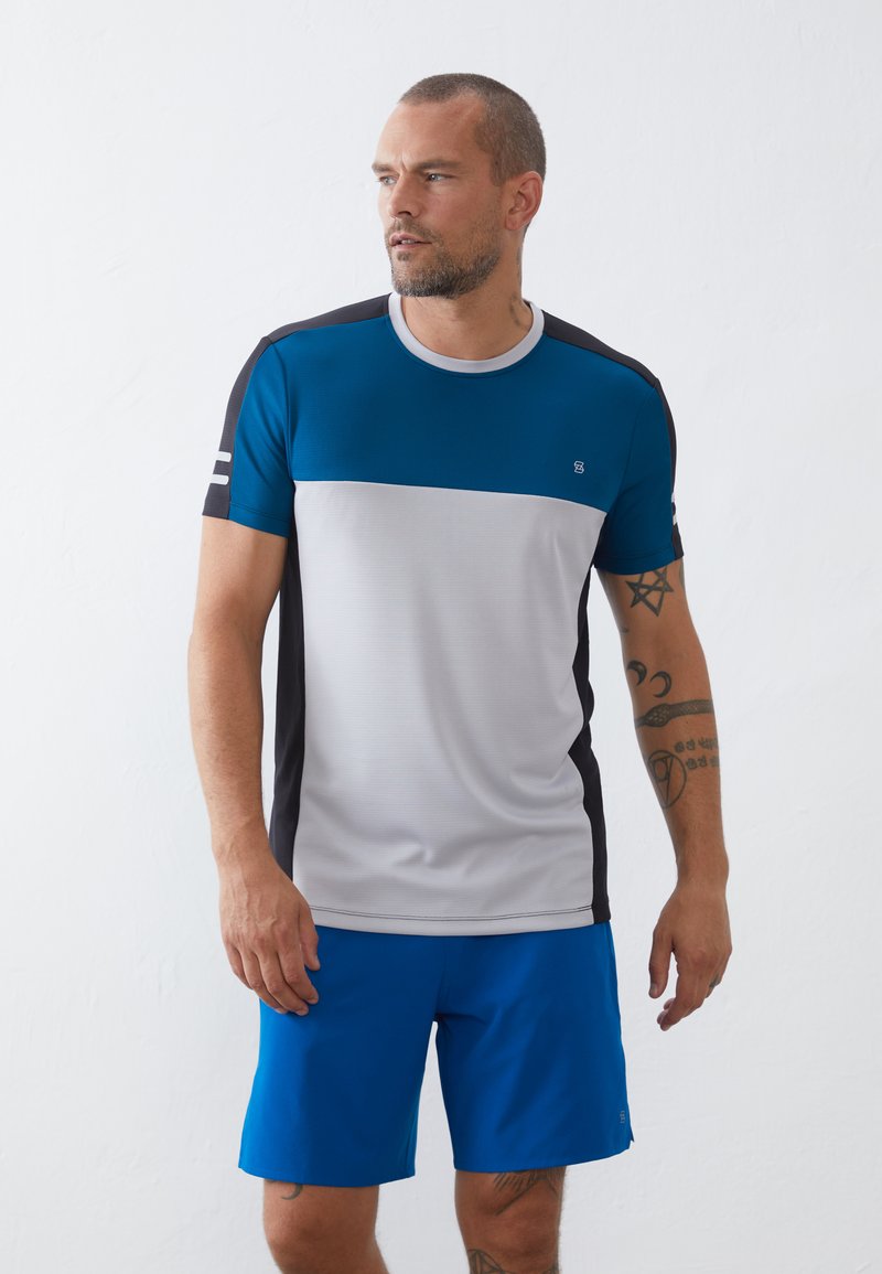 T-shirt masculino desportivo nas cores azul, cinza e preto, com decote redondo e mangas curtas. Textura suave com um design subtil. Combinado com calções azuis.