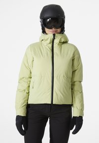 Hellgrünes, isoliertes Pufferjacke mit Kapuze, ausgestattet mit einem Frontreißverschluss, elastischen Bündchen und seitlichen Reißverschlusstaschen. Schwarze Skihandschuhe getragen.