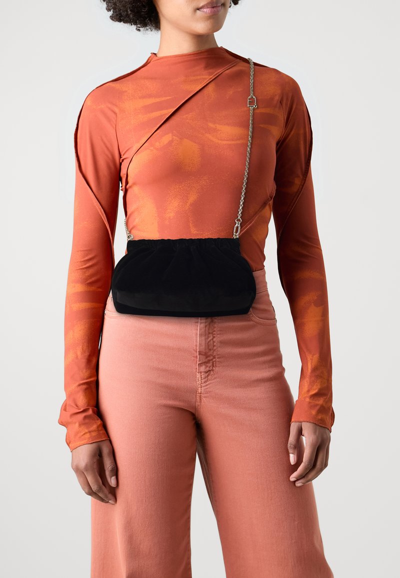 Saco de veludo preto com topo franzido e alça de corrente prateada, usado sobre uma blusa de manga longa laranja e calças rosa claro de cintura alta.