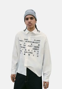 Camicia bianca a maniche lunghe con testo nero di varie dimensioni, caratterizzata da un colletto classico e chiusura con bottoni. Indossata con jeans scuri.
