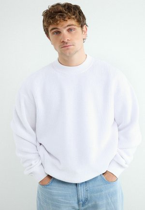 Mladý muž s kučeravými hnedými vlasmi, ktorý má na sebe biely pletený sveter a svetlomodré džínsy, ruky v vreckách, neutrálny výraz, biele pozadie.