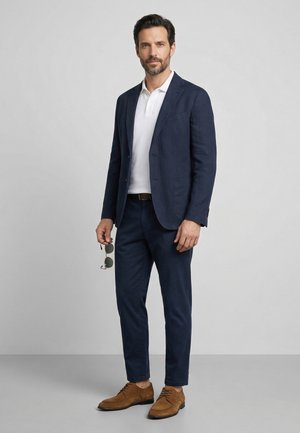 Man standing wearing navy blazer and pants, white polo shirt, brown dress shoes, holding sunglasses in his right hand.