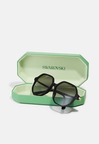 Ochelari de soare negri, în formă de octogon, cu lentile degradé, plasați într-un etui verde în formă de octogon. Etui-ul are o finisare texturată și logo-ul „Swarovski” embosat.