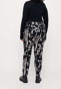 Žena oblečená v kalhotách s abstraktním vzorem v černé, bílé a fialové barvě, s černým dlouhým rukávem a černými botami, stojící na pozadí v jednobarevné bílé.