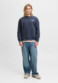 Sudadera azul marino con cuello redondo, puños acanalados y dobladillo. Llevada sobre una camiseta de color claro, combinada con unos vaqueros de tiro bajo y descoloridos en azul, y zapatos color tostado.