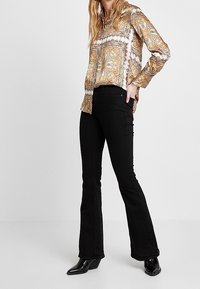 Blus i guld och vitt mönster med långa ärmar, i kombination med svarta utsvängda jeans och svarta klackboots. Slät tyg, skräddarsydd passform.