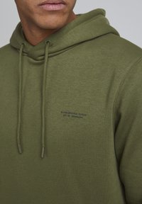 Hoodie vert olive en tissu doux, doté de cordons de serrage et d'un petit logo imprimé sur la poitrine. Texture lisse et coupe décontractée.