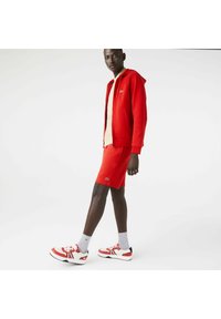 Punainen huppari, jossa on yhteensopivat shortsit, kerroksittain vaaleanbeige paitapuseron päällä. Jalkineissa on punaisia, valkoisia ja mustia yksityiskohtia. Harmaat sukat näkyvät.