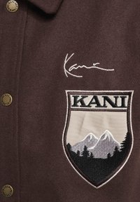 Hnedý vlnený blendový kabát s náprsním nášivkom s horami, stromami a textom "KANI"; obsahuje vyšívaný podpis a kovové gombíkové akcenty.
