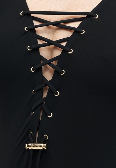 Κοντινό πλάνο μαύρου ενδύματος με μαύρα κορδόνια που περνούν μέσα από χρυσά περικάρπια, με χρυσή αγκράφα με το λογότυπο στο κάτω μέρος.