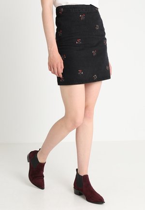 Mujer que lleva una minifalda negra con pequeños bordados florales rojos y botas de ante burdeos hasta el tobillo, de pie contra un fondo claro y uniforme.