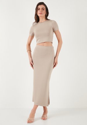 Bež cropped top i usklađena maksi suknja od mekog, rastegljivog materijala. Suknja ima bočni prorez. Model nosi bež sandale sa trakama.
