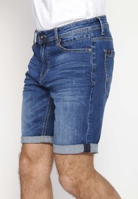 Pantalones cortos de denim en azul oscuro con un ligero desgastado. Presentan un dobladillo enrollado y un diseño de cinco bolsillos con costuras contrastantes y un cierre de botón.