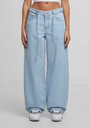 Persoon in ruime lichtblauwe denimjeans met een trekkoord in de taille en witte sneakers op een effen grijze achtergrond.