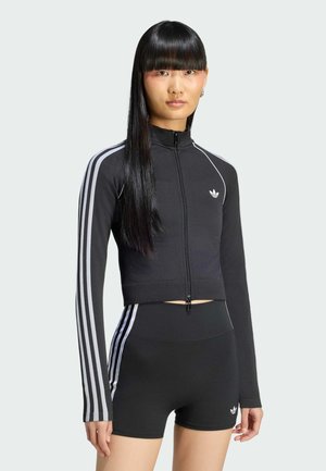 Jeune femme aux longs cheveux noirs portant une veste zippée noire Adidas et des shorts noirs assortis avec des bandes blanches.