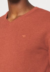 V-ringad tröja i rostfärg, tillverkad av ett mjukt stickat material. Har en diskret logopatch på bröstet samt ribbning runt halsringningen.