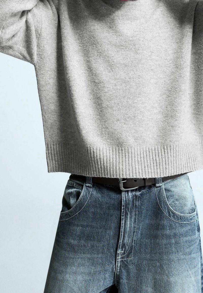 Personne portant un pull gris clair à manches longues, rentré dans un jean en denim bleu avec une ceinture noire, debout devant un fond clair.