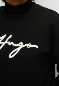 Schwarzer Sweatshirth mit einer großen, weißen gestickten Unterschrift "Hugo" auf der Vorderseite. Gerippter Rundhalsausschnitt und weicher Stofflack.