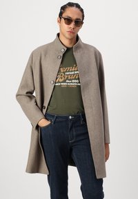 Ανοιχτό καφέ μάλλινο παλτό με ψηλό γιακά και τρία κουμπιά, φορεμένο πάνω από ένα πράσινο t-shirt με γραφικά και σκούρα μπλε τζιν. Τα γυαλιά ηλίου ολοκληρώνουν την εμφάνιση.