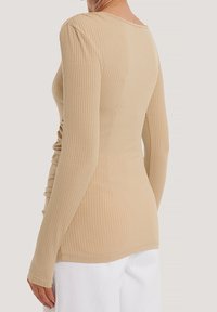 Haut beige à manches longues à côtes, coupe ajustée, avec une texture subtile et un col rond. Le tissu présente une finition lisse. Aucun élément métallique visible.