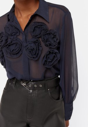 Granatowa prześwitująca bluzka z trójwymiarowymi kwiatami z tkaniny na przodzie, długie rękawy zakończone mankietami na guziki, w połączeniu z czarnymi skórzanymi spodniami z wysokim stanem i paskiem.