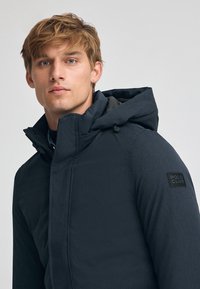 Giacca con cappuccio di un blu scuro, realizzata in tessuto testurizzato, con una patch nera "POLO CLUB" sulla manica e chiusura frontale con zip.