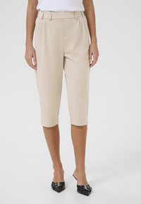 Beige, viistoreijalliset housut, joissa on tasainen etuosa, sivutaskut ja kääntöreunat, valmistettu sileästä kankaasta. Yhdistetty teräväkärkisiin mustiin kenkiin.