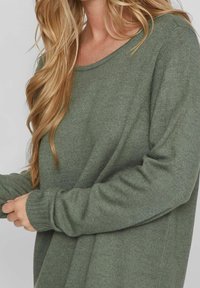 Pull en maille verte avec une coupe décontractée, un col large et des poignets côtelés. Le matériau est doux avec une texture subtile.