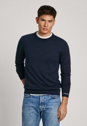 Marineblauer Strickpullover mit Rundhalsausschnitt und gerippten Bündchen, unterlegt mit einem weißen Shirt. Dezentes Logodetail am Ärmel. Kombiniert mit blauen Jeans.