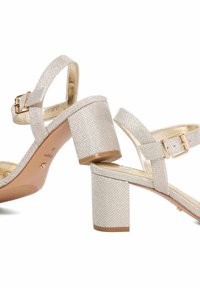 Sandales à talons hauts beiges texturées avec un talon bloc, une sangle ajustable à la cheville et des accents de boucle métallique. Doublure intérieure lisse.