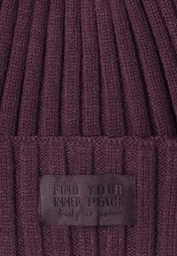 Strikket beanie i mørk lilla med ribbet tekstur. Sort etiket syet på fronten viser "FIND YOUR INNER PEACE" i præget tekst.