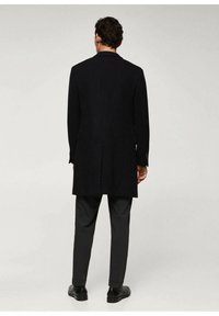 Manteau en laine noire à coupe droite, dos boutonné et coupe ajustée, associé à un pantalon gris foncé et des chaussures noires.