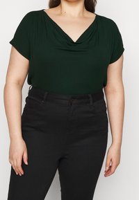 Mörkgrön, draperad, kortärmad topp med V-ringning. Bärs med högmidjade svarta jeans som har fickor och ett knappstängning.