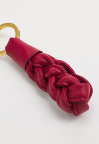 Porte-clés en cuir rouge avec un design tressé, texture lisse et anneau en métal doré. Le cuir présente un léger éclat et une forme flexible.