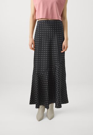 Jupe maxi noire à pois avec un ourlet évasé, fabriquée dans un tissu lisse. Portée avec un crop top rose et des bottines beige à talons pointus.