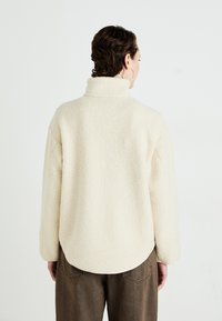 Person with short brown hair faces away, wearing cream-colored fleece jacket with high collar and brown pants.