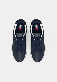 Baskets bleu marine en matériau synthétique lisse, semelles en caoutchouc blanc, et une étiquette de logo avec des rayures verticales rouges, blanches et bleues.