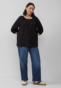 Zwarte, gestructureerde blouse met een ronde hals en geplooide details, gecombineerd met blauwe denim jeans en beige sneakers.
