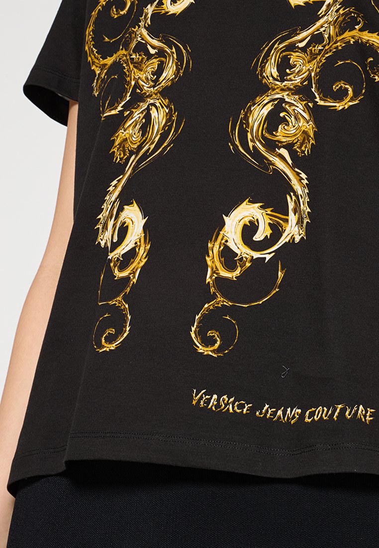 T-shirt noir avec un motif imprimé doré tourbillonnant. Le texte "VERSACE JEANS COUTURE" est brodé près de l'ourlet. Tissu doux.