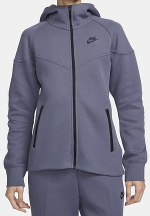 Kobieta nosząca fioletową bluzę Nike z suwakiem na całej długości, kapturem, dwoma zapinanymi na suwak kieszeniami po bokach i dopasowanymi fioletowymi spodniami dresowymi Nike.