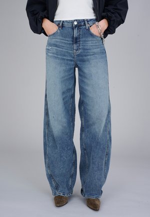 Osoba w szerokich, wyblakłych jeansach z szerokimi nogawkami, z rękami w kieszeniach, z łańcuszkowym dodatkiem, brązowych, spiczastych butach, białym topie i czarnej kurtce.