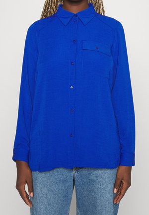 Persoon draagt een felblauw overhemd met lange mouwen en borstzak, gecombineerd met lichtblauwe jeans, staand tegen een effen achtergrond.